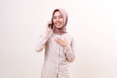 Relieved young asian muslim girl holding her chest while talking on the phone. Isolated on white background