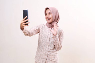 Happy smiling expression of young asian muslim girl while doing selfie with her cell phone. Isolated on white background