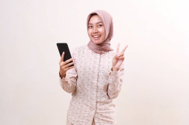 Excited young asian muslim girl standing showing two fingers while holding a cell phone. Isolated on white