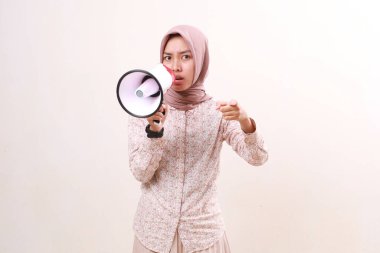 Angry young asian muslim woman using megaphone while pointing at you or camera. Isolated on white