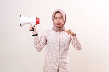 Unhappy young asian muslim woman using megaphone while pointing at you or camera. Isolated on white