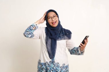 Neşeli Asyalı yaşlı Müslüman kadın elinde cep telefonuyla mutlu bir yüz ifadesiyle ayakta duruyor.