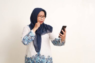 Kafası karışmış Asyalı yaşlı Müslüman kadın elinde cep telefonuyla dikiliyor.