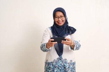 Mutlu Asyalı yaşlı Müslüman kadın akıllı telefonuyla video izlerken ayakta duruyor.