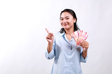 Smiling young modern asian woman, pointing at empty space, holding cash money rupiah, standing over white background looking at the camera clipart