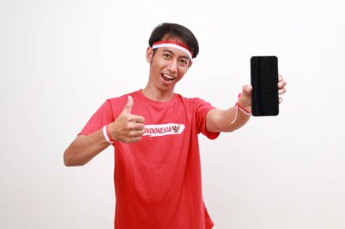 Endonezya 'nın bağımsızlık gününü kutlayan kırmızı beyaz kurdeleli mutlu Asyalı çocuk cep telefonu tutarken başparmağını kaldırıyor.