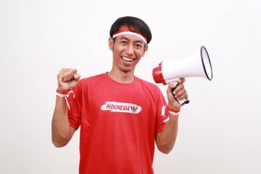 Asian man celebrating Indonesia Independence day, holding megaphone while clenching hand. Isolated on white clipart