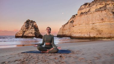 Gün batımında manzaralı bir plajda Lotus Pozu pratiği yapan kadın..