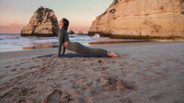 Gün batımında yüzükoyun plaja bakan köpek pozu veren kadın omurgasını geriyor ve göğsünü açıyor..
