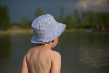 Sırtı suyun kenarında bir şapkanın içinde duran ve mesafeye bakan bir çocuk. Dört yaşında bir çocuk.