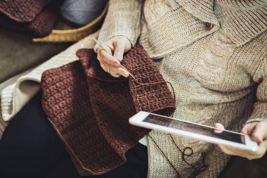 El yapımı ev yapımı hobi evde kalma konsepti mutlu Asyalı yaşlı kadın evdeki oturma odasındaki kanepede kahverengi fular ve tablet örüyor.