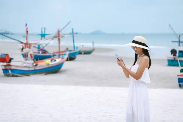 タイの夏の海釣りボートの背景に対してビーチで携帯電話を使用して白いドレスを持つ若い旅行アジアの女性 ストック画像