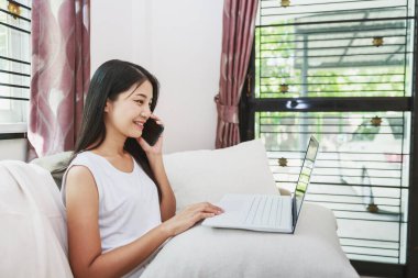 Evden ve internetten alışveriş kavramından mutlu Asyalı kadın cep telefonu ve dizüstü bilgisayarını evdeki oturma odasındaki koltukta dizüstü bilgisayarla kullanıyor.