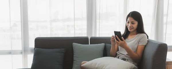 自宅やオンラインショッピングコンセプトからのレジャー ハッピーアジアの女性はリラックスして自宅のリビングルームでソファーで携帯電話を使用 ストックフォト