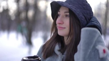 Genç, zayıf, siyah saçlı, kahverengi gözlü, kağıt bardaktan kahve içen, açık havada gülümseyen bir kadın. Rahatlamış beyaz kadın portresi. Kış tatilinin tadını çıkarıyor.