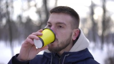 Yakından kendine güvenen esmer, sakallı, kahverengi gözlü, kağıt bardaktan kahve içen, kış parkında dikilip bakan adam. Beyaz adamın portresi. Açık havada eğleniyor.