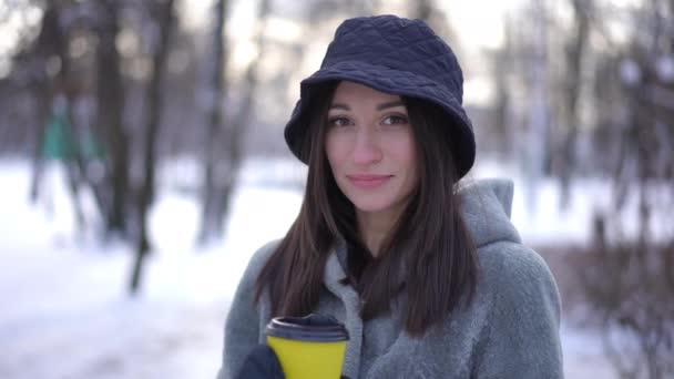 屋外の使い捨てカップからコーヒーを飲むカメラを見て魅力的な若い女性を笑顔 冬の公園で熱い飲み物を楽しむスローモーションでポーズをとるスリムな美しい白人女性の肖像画 — ストック動画