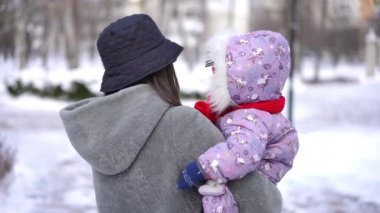 Kış parkında küçük tatlı bir kızla yürürken gülümseyen mutlu genç bir kadının fotoğrafını çekiyorum. Beyaz tenli güzel anne, çocuğu elinde tutuyor. Soğuk bir günde ağır çekimde geziyor.