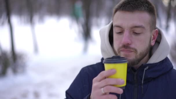 雪の多い冬の公園でコーヒーを飲みながらカメラを見ている若い白人男性 真剣に自信を持って積極的な男の肖像画は屋外でスローモーションでポーズ — ストック動画