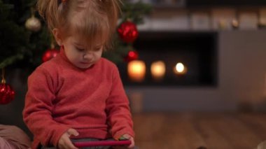 Sevimli küçük kız hapşırıyor gülümsüyor gözlerini kaçırıyor oturma odasında şömineyle akıllı telefonuyla oynuyor. Noel arifesinde evde solda oturan beyaz bir çocuğun portresi.