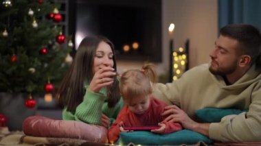 Mutlu genç ebeveynler, akıllı telefondan küçük kızlarını öperek çizgi film izliyorlar. Olumlu gülümseyen Kafkasyalı karı koca evde güzel bir kızla Noel tatilinin tadını çıkarıyor.