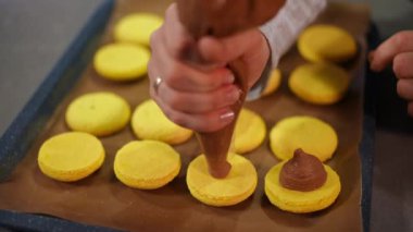 Yavaş çekimde sarı Fransız kurabiyelerinin üzerine aşçı şırıngasından çikolata sıkıyorum. Tanımlanamayan beyaz kadın mutfaktaki tatlı hamur işlerini pişiriyor.