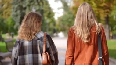 Canlı kamera, parkta yürüyen ve gülümseyen iki genç bayanı takip ediyor. Beyazların güz gününde dışarıda gezinip sohbet ederken orta açıdan çekilmiş bir fotoğrafı.