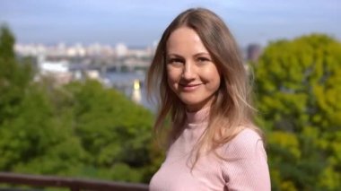 Genç, pozitif bir kadın kameraya bakıyor ve açık havada ağır çekimde gülümsüyor. Bulanık şehir ve nehrin arka planında poz veren kendinden emin mutlu beyaz kadın portresi.