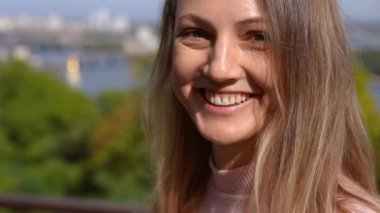 Yavaş çekimde gülümseyen genç ve çekici bir kadının yakın plan portresi. Park nehrinin arka planında poz veren Kafkasyalı mutlu kadının vesikalığı ve bulanık şehir
