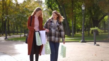 Alışveriş çantalı iki mutlu kadın güneşli parkta yürüyüp giderken konuşuyorlar. Tatmin olmuş genç, güzel beyaz alışverişçilerin portresi Kara Cuma 'da ağır çekimde dışarıda geziniyor.