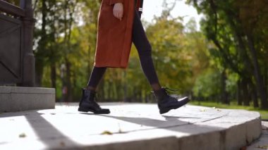 Tanımlanamayan zayıf, stil sahibi genç bir kadının parkta taş merdivenlerden yavaşça inişini gösteren bir yan görüntü. Kendine güvenen beyaz kadın gün ışığında dışarıda geziniyor.