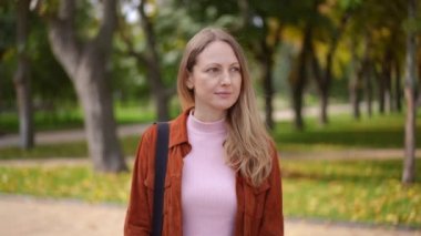 Canlı kamera, düşünceli, kendine güvenen genç bir kadının park alanında durup gülümseyen kameraya bakışına odaklan. Ağır çekimde poz veren pozitif beyaz kadın portresi.