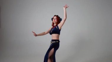 Gri arka planda dans eden zayıf, esnek, kendine güvenen bir kadın. Geniş açılı portre, çıplak ayaklı güzel Kafkasyalı kızıl saçlı dansçı Orta Doğu dansı yapıyor.