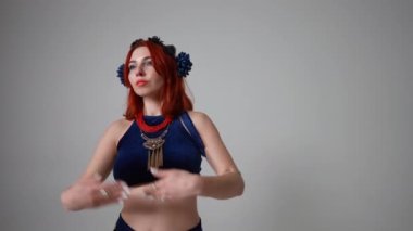 Genç, sıska bir kadın ağır çekimde dans ederek el kaldırıyor. Kızıl saçlı beyaz kadının portresi gri arka planda göbek dansı yapıyor.