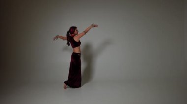 Kolunda kanat dövmesi olan zayıf bir kadın gri arka planda ağır çekimde el kaldırıyor. Orta Doğu dansında dans eden beyaz yetenekli dansçı.