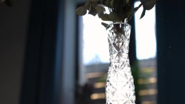Yakın plan kristal vazo. İçerideki masada güneş ışığı altında çiçekler var. Güneş ışınlarında saydam konteynır. Yavaş çekim