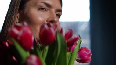 Mutlu, kendine güvenen güzel bir kadının yakından çekilmiş bir portresi. Gün ışığında yavaş çekimde çiçekleri koklarken başka tarafa bakıyor. Beyaz tenli genç bayan solda ayakta duruyor.