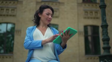Orta ölçekli, zarif takım elbiseli, beyaz bir kadının dijital tablette internette sörf yapıp kentteki bankta otururken çekilmiş resmi. Kendine güvenen çekici iş kadını yavaş çekimde internetten mesajlaşıyor.
