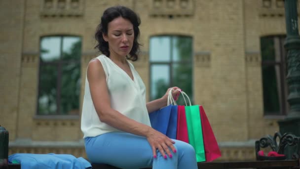 疲れた妊婦がショッピングバッグでベンチに座って痛みを伴う足を撫でて腹を愛撫 街の美しい白人のブルネットの女性の肖像画 スローモーション — ストック動画