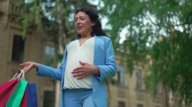 Kendine güvenen güzel bir kadının hamile karnını okşayıp alışveriş torbalarını yavaş çekimde kaldırdığı orta boy bir fotoğraf. Mutlu Kafkasyalı zarif bayan Kara Cuma satışlarında gülümsüyor ve başka tarafa bakıyor.