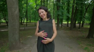 Park sokağında duran kameraya yüksek sesle gülen neşeli hamile kadının ön manzara portresi. Kafkasyalı mutlu güzel bayan dışarıda ağır çekimde poz veriyor.