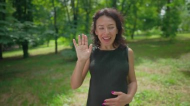 Neşeli hamile kadın, güneşli yeşil park çayırında kameraya bakarak konuşuyor. Siyah elbiseli neşeli beyaz kadının portresi ağır çekimde sallanan blog yazarken
