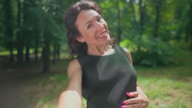 Kendine güveni tam hamile blogcunun kameraya bakıp suratını buruşturan selfie POV 'u. Parkta ağır çekimde güneşin altında poz veren neşeli beyaz kadın portresi