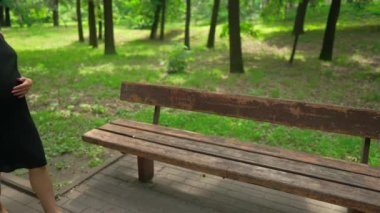 Sunny Park 'taki ahşap bankta oturan hamile bir kadın gülümsüyor. Siyah elbiseli zarif bir bayanın portresi bahar yaz günü açık havada sükunetin tadını çıkarıyor. Yavaş çekim