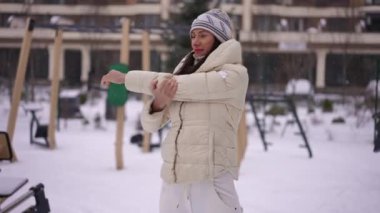 Kafkasyalı gülen sporcu kadın açık havada karlı bir kış günü ısınıyor. Kendine güvenen genç, zayıf, kaslarını esneten, spor yapan bir kadının orta boy portresi.