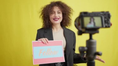 Gülümseyen ve konuşan genç bir kadın Follow plaketini ve bulanık video kamera görüntülerini gösteriyor. Kendine güvenen güzel beyaz kadın blog yazarının portresi sarı arka planda hikayeler kaydediyor.