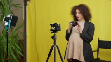 Kamera ve ışık standı olan tripodlu bulanık hamile kadın sarı arka planda film blogu ile konuşuyor. Genç Kafkas beklenen blogcu reklam ekipmanları içeride vlog kaydediyor
