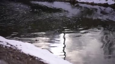 Winter sunshine reflecting in cold river water with snow on shore. Close-up calm stream outdoors on autumn spring day