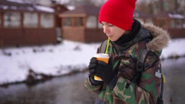 Relaxed Caucasian teen boy drinking coffee walking on sunny winter day along river shore. Portrait of carefree confident teenager strolling outdoors after school thinking. Solitude and lifestyle