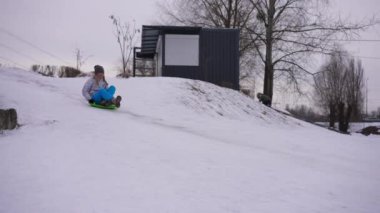 Wide shot slope with white snow and cheerful teen girl passing on green sled. Happy excited Caucasian charming teenager enjoying winter leisure outdoors on cloudy day. Slow motion